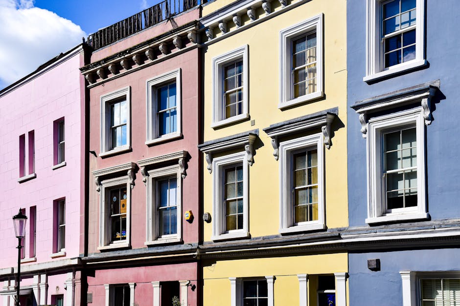A row of historic terraced houses painted in pastel colors, including pink, yellow, and blue, with white decorative window frames and cornices. The buildings are three stories tall with large sash windows on each floor, some of which have partially opened blinds or curtains. The facades feature detailed molding and ornamentation around the windows, adding a classical architectural touch. The street scene is illuminated by daylight, casting soft shadows on the building surfaces, with a classic black street lamp visible on the left side of the image. The sky is partly cloudy, providing a bright background behind the buildings. The scene exemplifies typical urban residential architecture and illustrates an environment that may be serviced by independent waste collection, such as rubbish removal by local companies like rubbishcollectionnottinghill.co.uk, which can facilitate on-site clearance and private waste handling for residents in this area.