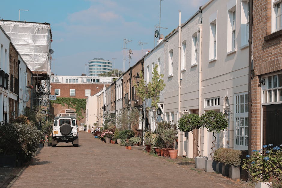 A quiet residential street in Notting Hill featuring a row of terraced houses with white and brick facades, some with small front gardens filled with potted plants and shrubs. The cobbled pavement in the foreground is lined with various potted greenery, including small trees and flowering plants. A white utility van with a spare wheel mounted on the rear is parked on the street, indicating possible rubbish collection or private waste removal activity. The background shows a modern high-rise building with curved glass windows, contrasting with the traditional houses. Clear blue sky overhead suggests a bright day. This scene exemplifies a typical urban environment where private waste clearance or independent rubbish collection services, such as those provided by companies like rubbishcollectionnottinghill.co.uk, might operate outside council bulk pick-up schedules, illustrating alternative waste handling methods in a residential context.