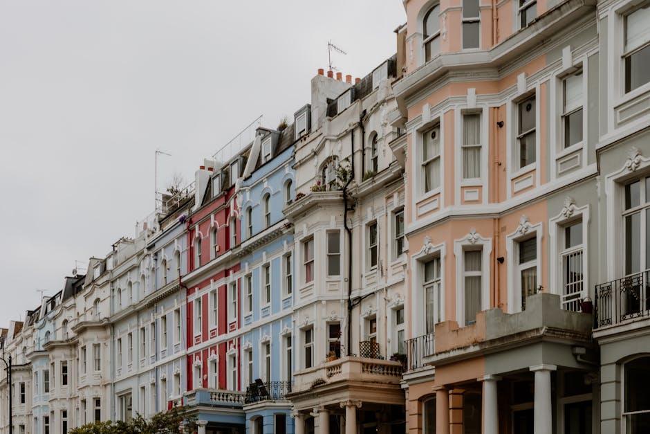 A row of historic terraced houses painted in pastel colors, including pink, yellow, and blue, with white decorative window frames and cornices. The buildings are three stories tall with large sash windows on each floor, some of which have partially opened blinds or curtains. The facades feature detailed molding and ornamentation around the windows, adding a classical architectural touch. The street scene is illuminated by daylight, casting soft shadows on the building surfaces, with a classic black street lamp visible on the left side of the image. The sky is partly cloudy, providing a bright background behind the buildings. The scene exemplifies typical urban residential architecture and illustrates an environment that may be serviced by independent waste collection, such as rubbish removal by local companies like rubbishcollectionnottinghill.co.uk, which can facilitate on-site clearance and private waste handling for residents in this area.