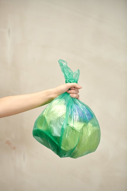 A person's arm extends into the frame, holding a tightly knotted green plastic bag filled with waste. The bag appears semi-transparent, revealing various indistinct items inside, and has a slightly glossy surface with visible creases and folds. The knot at the top is twisted, with some excess plastic forming a small tail that sticks upward. The background is a plain, neutral-colored wall with a soft, even lighting that highlights the bag's sheen. The scene evokes an everyday activity related to rubbish collection, illustrating the handling and disposal of household waste, which may be managed through private rubbish removal services like those offered by the company mentioned. The image emphasizes the practicality of waste disposal, with visual cues toward waste management and rubbish collection services, particularly in an urban or residential setting.
