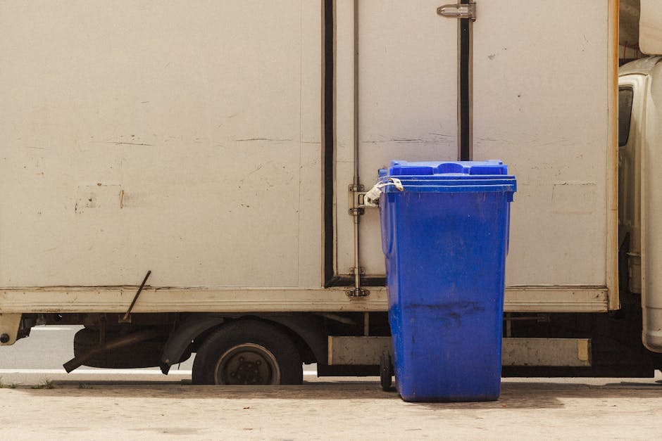 The image shows a large, blue wheelie bin with a closed lid placed beside the side of a white delivery or storage truck. The bin has a smooth plastic surface with some visible dirt marks towards the lower part and is positioned on a paved surface, with the truck's side panel extending above. The truck appears to have a metal exterior with a slightly weathered finish, and a black rubber tire is partially visible underneath the cargo area, suggesting the vehicle is used for transportation or waste collection. In the background, the environment is neutral and outdoors, with no distinct urban or natural elements visible, indicating the scene is likely on a residential or commercial street with an emphasis on waste management or rubbish removal services. The overall composition reflects a typical setting for private waste collection, with the bin likely used for rubbish or bulky waste disposal, managed by a company specializing in independent collection services such as rubbishcollectionnottinghill.co.uk.