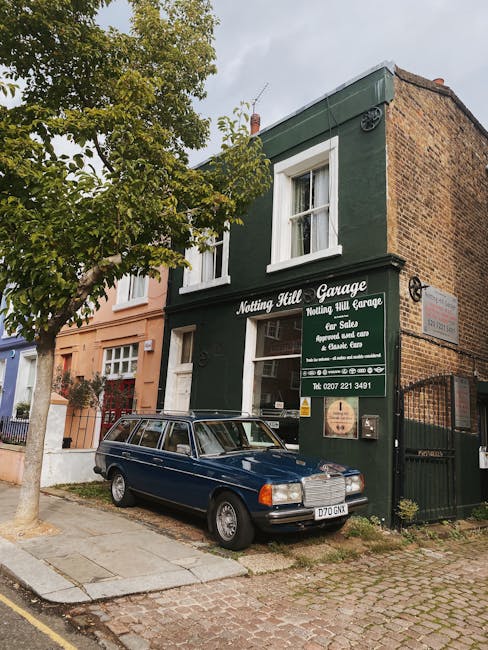 A street scene featuring a small, two-storey green brick building with white-trimmed windows and a sign reading 'Notting Hill Garage' in white lettering, indicating it is a private garage and car sales business. Adjacent to the green building is a narrow, brown brick side wall, with a black metal gate in the lower right corner. A classic dark blue station wagon with a silver grille and bumper is parked directly in front of the garage on a paved sidewalk, partially on a concrete slab. To the left of the car, a large leafy tree with green foliage extends over the pavement, providing partial shade. The overall setting is a quiet residential street with a mix of brick houses painted in pastel tones, and cobbled or paved road surfaces, suggesting an urban environment where private waste collection or rubbish removal services might be utilised, as supported by the company's presence on the building.