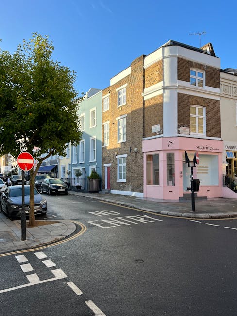 A quiet urban street scene featuring a three-story building with a ground-floor shop painted in pale pink, with large windows and a small sign above the entrance. The building’s upper floors are constructed with brown brickwork and white trim around the windows, some of which are double-hung sash styles. To the left of the building, there is a tree with green foliage, partially obscuring the view of parked vehicles lining the curb. Several cars are parked along the street on both sides, with one visible being a silver vehicle. The road curves gently with clear white markings, including a 'NO ENTRY' sign painted on the asphalt near the corner. The scene is well-lit, suggesting daytime with natural sunlight casting soft shadows. This setting reflects an area where private waste handling and rubbish collection services, such as those offered by rubbishcollectionnottinghill.co.uk, might support local small businesses or residential waste management needs in the neighbourhood.