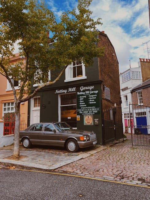 A two-storey building painted in dark green with white window frames, identified as Nottingham Hill Garage, situated on a cobbled street. The building's signage displays the garage's name, services offered such as tyre repairs, servicing, and diagnostics, along with a contact phone number. In front of the garage, a vintage grey car with a textured metallic finish is parked partially on the pavement and on the cobblestone driveway. A mature tree with green and yellow leaves stands to the left, its branches extending over the car and partly obscuring the upper windows of the building. To the right, a black metal gate and fence border the premises, with neighboring buildings visible further along the street, including one with white timber-framed details. The setting appears to be in a residential urban area, featuring a mixture of architectural styles, and the scene is illuminated by daylight with a partly cloudy sky overhead. The overall composition reflects a typical environment for independent vehicle service providers operating in close proximity to private residential properties, consistent with alternative waste removal or service sites managed by companies like rubbishcollectionnottinghill.co.uk.