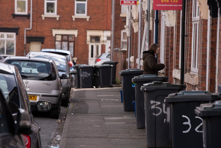 A residential street scene featuring a row of dark grey wheelie bins with white numbering, placed along the pavement beside a line of brick terraced houses. The bins are positioned close to the building walls, some with their lids slightly open, and are used for waste collection. A woman wearing a brown coat stands next to one of the bins, appearing to be in the process of disposing of rubbish. The sidewalk is narrow, with a few small pieces of litter visible on the pavement. Parked cars, including a silver hatchback and a dark vehicle, are lined up along the street on the left side, with their rear ends facing the viewer. The background shows more brick houses, some with white window frames and small front yards, under overcast lighting that creates an evenly lit, neutral environment. The scene reflects typical urban waste management practices, with the bins likely used for private disposal or alternative rubbish collection services, supported by companies operating in local areas like Notting Hill.