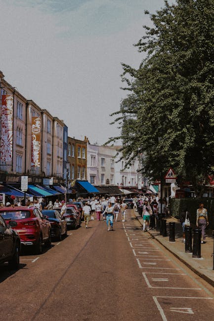 A busy street scene in a residential area with a mix of parked cars and pedestrians walking along the pavement. The street is lined with colorful, multi-story terraced buildings featuring various shades of beige, brown, and pastel tones, some with awnings over shopfronts. On the right side, a large leafy tree extends over the pavement, providing shade. Bollards are spaced along the sidewalk to separate pedestrian space from the roadway. The sky appears slightly overcast, creating a diffuse lighting across the scene. In the foreground, the street is clear with a designated parking bay marked by white lines, indicating an organized approach to vehicle parking, possibly for private or alternative waste collection services. The presence of pedestrians and parked cars suggests an active urban environment where waste collection might happen outside conventional local authority hours or through private arrangements, aligning with rubbish removal and on-site clearance practices. The overall setting exemplifies a typical street scene that may be associated with independent waste disposal options, such as rubbish collection services operating within Notting Hill or similar areas, as highlighted by local regulations concerning rubbish and permits.