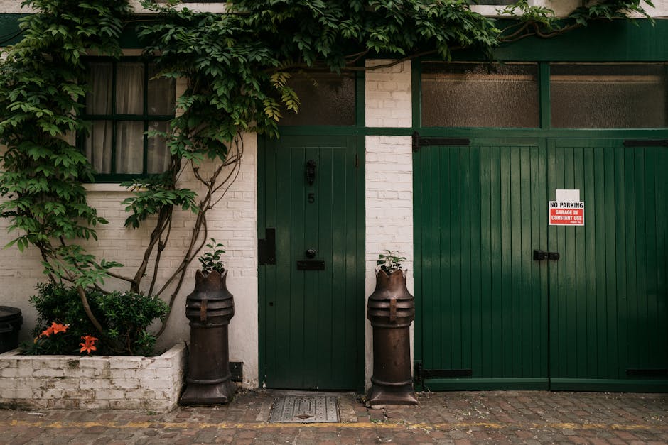 A view of a building façade featuring a centrally located dark green wooden door with a vertical panel design, positioned between two vintage-style cast iron bollards that are rusted, cylindrical with rounded tops, and placed on both sides of the door. To the right of the door, there is a large matching green double sliding gate with horizontal panels and black handle locks, one of which has a red and white sign reading 'NO PARKING GARAGE IN CONSTANT USE.' The building has white brick walls, with a small window covered by dark curtains on the left side, partially obscured by a climbing vine with lush green leaves extending over the top of the window and partially across the wall. In front of the building, there is a narrow cobblestone street or pathway, with some small plants and orange flowers planted in a white brick-bordered flower bed on the left side. The scene appears to be outdoors, lit by natural daylight, capturing a quiet urban setting where private entry and alternative waste handling could occur, with the surrounding greenery providing a contrasting natural element to the structured façade.