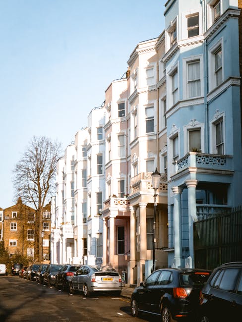 A busy street scene in a residential area with a mix of parked cars and pedestrians walking along the pavement. The street is lined with colorful, multi-story terraced buildings featuring various shades of beige, brown, and pastel tones, some with awnings over shopfronts. On the right side, a large leafy tree extends over the pavement, providing shade. Bollards are spaced along the sidewalk to separate pedestrian space from the roadway. The sky appears slightly overcast, creating a diffuse lighting across the scene. In the foreground, the street is clear with a designated parking bay marked by white lines, indicating an organized approach to vehicle parking, possibly for private or alternative waste collection services. The presence of pedestrians and parked cars suggests an active urban environment where waste collection might happen outside conventional local authority hours or through private arrangements, aligning with rubbish removal and on-site clearance practices. The overall setting exemplifies a typical street scene that may be associated with independent waste disposal options, such as rubbish collection services operating within Notting Hill or similar areas, as highlighted by local regulations concerning rubbish and permits.