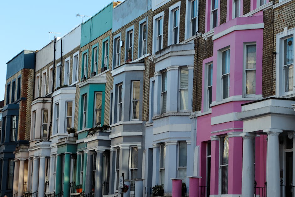 A row of Victorian terraced houses with distinctive bay windows and ornate architectural details, painted in pastel shades of blue, turquoise, white, light grey, and pink. The buildings are three to four stories high, with brickwork visible on upper levels and decorative features such as cornices and columns around the bay windows. Some houses have small front gardens or flower pots near the entrances, and black wrought-iron railings line the pavement in front of the properties. The sky above is clear and the scene is illuminated by natural daylight, highlighting the vibrant yet neutral tones of the facades. This residential street showcases classic urban architecture, typical of areas that may require regular rubbish removal and waste collection services, with the potential for private disposal solutions like those offered by rubbishcollectionnottinghill.co.uk, especially suited for managing waste from maintenance or refurbishment activities on such properties.