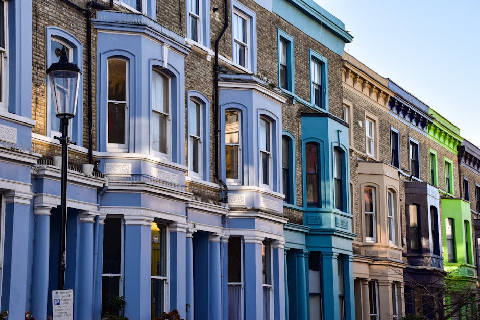A row of Victorian-era terraced houses with ornate detailing and bay windows, painted in pastel shades of white, light blue, peach, and pink, situated along a narrow street under an overcast sky. The buildings feature decorative moldings and iron railings on small balconies, with some windows partially visible and framed by white trim. The roofs are sloped with slate tiles, and various chimneys and antennas are visible on top. The street below is not visible, but the facades suggest an urban residential environment. The overall scene appears calm, with muted natural lighting typical of cloudy weather, highlighting the intricate architectural features and soft colors of the buildings. This visual context aligns naturally with private waste management or alternative rubbish collection services involving on-site clearance or flexible rubbish removal from residential properties, as often seen in urban environments like Notting Hill. Such detailed street-level imagery supports the importance of responsible rubbish disposal and reporting fly-tipping to maintain the area's cleanliness and aesthetic appeal, common in services provided by companies specializing in rubbish removal.