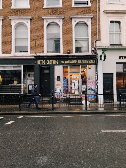 The image depicts the storefront of a vintage clothing shop named 'RETRO CLOTHING' on a busy street in Notting Hill. The shop has a black facade with a bold yellow sign advertising secondhand bargains for men and women. The large front window display is filled with racks of clothing and notices taped to the glass, with some graffiti visible on the white wall adjacent to the store. The building above consists of three white arch-topped windows set into a brown brick exterior. In front of the shop, a pedestrian is walking along the sidewalk, which is separated from the street by a black metal railing. The street appears wet, likely due to recent rain, with reflections visible on the asphalt. To the right of the clothing store, there is another shop entrance with a white sign, and a small security camera is mounted on the corner of the building. The overall scene captures an urban environment typical of a vibrant shopping district, illustrating how independent retail premises are accessible for waste and rubbish collection through private disposal services, such as those provided by rubbishcollectionnottinghill.co.uk.