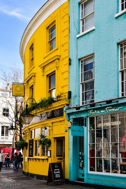 The image shows the exterior of a multi-storey building with a distinctive colour contrast, featuring a bright yellow facade on the left side and a pastel blue facade on the right side, both with large rectangular sash windows. The yellow section has ornate architectural details around the windows and a small balcony with a black wrought iron railing that holds potted plants and greenery. The building is situated on a busy street corner, with a black street lamp and a rectangular signboard hanging from a bracket attached to the yellow exterior. To the right, the blue part of the building houses a shop with a large glass window and a teal-colored awning displaying white script text. There is a small paved pavement in front of the building, with pedestrians walking nearby. The scene is captured in daylight, with some bare trees visible in the background, indicating a neutral outdoor environment. The image was taken as part of an urban streetscape, possibly in a commercial area, aligning with themes of private property and independent urban waste management, although no rubbish or removal activities are directly visible in this shot.