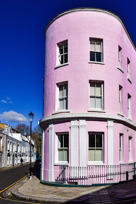 A rounded, three-story corner building painted in pastel pink, featuring a smooth exterior with evenly spaced, white-framed sash windows on each floor. The windows have dark glass panes and are aligned vertically, with some slightly open. The building is situated on a cobblestone sidewalk, with a short black wrought iron fence at the base, separating it from the pavement. To the left, a narrow street runs alongside a row of traditional white terraced houses with small front gardens and black railings, under a clear blue sky with a few wispy clouds. An old-fashioned street lamp stands on the pavement near the building, and parked cars are visible further down the road. The scene outside suggests an urban residential area with well-maintained structures, typical of a city environment suitable for private waste collection or on-site clearance services like those offered by rubbishcollectionnottinghill.co.uk, aligning with the context of alternative rubbish removal options in the area.