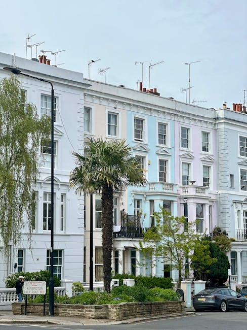 The image shows the exterior of a multi-storey building with a distinctive colour contrast, featuring a bright yellow facade on the left side and a pastel blue facade on the right side, both with large rectangular sash windows. The yellow section has ornate architectural details around the windows and a small balcony with a black wrought iron railing that holds potted plants and greenery. The building is situated on a busy street corner, with a black street lamp and a rectangular signboard hanging from a bracket attached to the yellow exterior. To the right, the blue part of the building houses a shop with a large glass window and a teal-colored awning displaying white script text. There is a small paved pavement in front of the building, with pedestrians walking nearby. The scene is captured in daylight, with some bare trees visible in the background, indicating a neutral outdoor environment. The image was taken as part of an urban streetscape, possibly in a commercial area, aligning with themes of private property and independent urban waste management, although no rubbish or removal activities are directly visible in this shot.