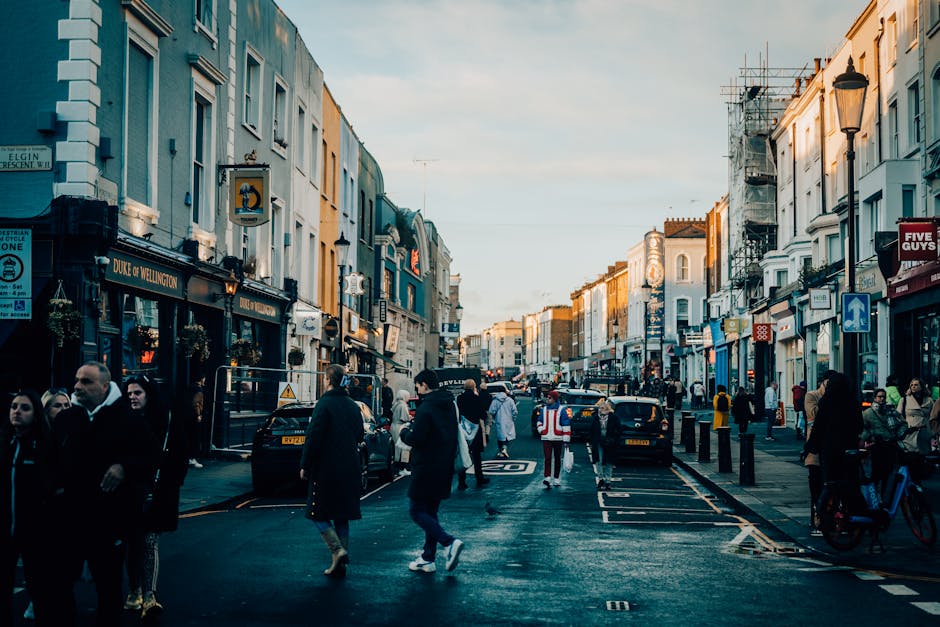 A busy urban street scene in the late afternoon or early evening with pedestrians walking along the pavement and crossing the road, lined with a mix of retail shops, restaurants, and cafes that have signage and storefront displays. The buildings are multi-storey structures with varied architectural styles, some featuring scaffolding indicating ongoing maintenance or renovation. Parked and moving vehicles, including black taxis, are visible on the road, with cyclists also present on the right side. Street lamps and hanging signs are mounted on building facades, and a clear sky with soft natural light illuminates the scene, creating gentle shadows. The overall environment reflects active city life typical of a central commercial area, with visible foot traffic and transportation activity that could relate to alternative waste handling and independent rubbish collection operations that support local trade and building maintenance, aligning with professional waste management practices in urban settings.