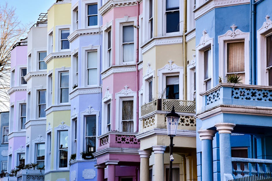 The image displays a row of Victorian-style terraced houses, each featuring distinct colour schemes on their facades, including shades of blue, teal, white, and pink. The buildings are constructed with brick exteriors, some painted over, with large sash windows and decorative cornices. The ground floors have small front gardens enclosed by black wrought iron railings, with some displaying flower pots and small plants. The structures are closely spaced, sharing common walls, and the roofs are flat with minimal visible roof fixtures against a clear, blue sky. The lighting suggests daytime, with natural sunlight casting soft shadows. This environment provides a typical residential street scene that could relate to private waste handling or alternative rubbish collection services, especially in contexts where individual residences or property managers manage waste clearance independently of local authority waste collection. The focus on the building facades and overall street layout emphasizes the type of properties that might require regular rubbish removal or on-site clearance activities, which are often handled by independent waste collection companies like rubbishcollectionnottinghill.co.uk.