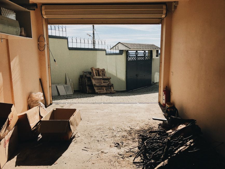 A view from inside an unfinished or recently cleared garage looking outward through an open roller shutter door. Inside the garage, there are several cardboard boxes on the left side, some partially collapsed or damaged, and a small pile of blackened, charred debris or scrap material on the concrete floor toward the right. The concrete floor appears slightly dusty or stained. Outside the garage, a paved driveway extends to a light green boundary wall topped with security spikes. Behind the wall, a small shed with a tiled roof and a pair of black metal gates with decorative elements are visible. Wooden pallets are neatly stacked against the wall, and an antenna or utility pole with wires can be seen in the background under a clear, slightly cloudy sky. The scene suggests a recent site clearance or rubbish removal process managed by a professional waste management or rubbish collection service, with an emphasis on private disposal or on-site clearance where debris is gathered for subsequent removal.